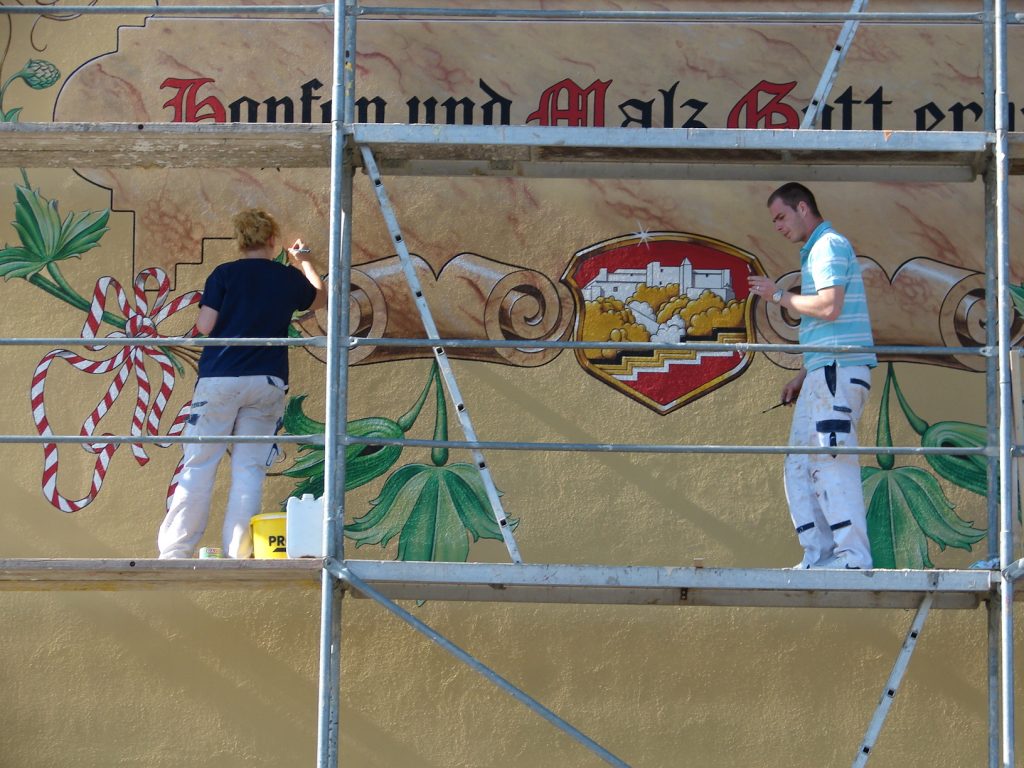 Malermeisterin Sebela-Stundner und ihr Mitarbeiter bei der Arbeit, stehend auf einem Baugerüst. Eine Fassade wird kunstvoll verziert.